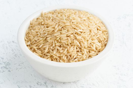 Brown rice in a white bowl on white background. Dried cereals in cup, vegan food, fodmap diet. Side view, close up.