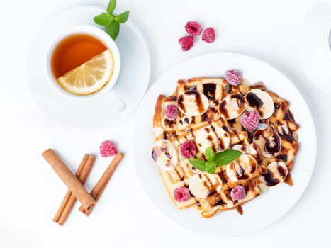 Belgian curd waffles with raspberries, banana, chocolate syrup. Breakfast with tea on white background, top view