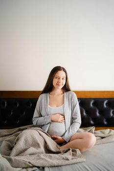 Pregnant woman sits on the bed and hugs her belly with her hands. High quality photo