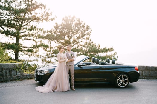 Bride and groom stand near the convertible on the road. High quality photo
