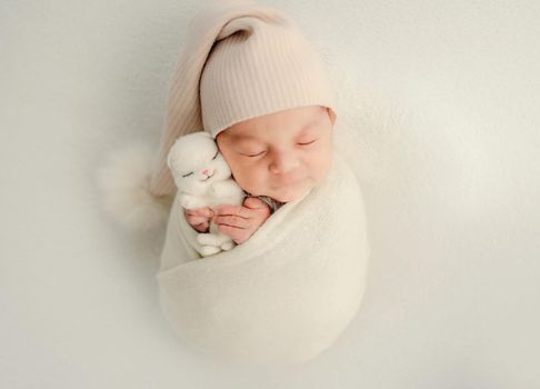 Newborn baby girl swaddled in white fabric holding cute bunny toy and sleeping. Adorable infant child kid napping wearing hat