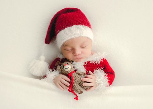 Newborn baby girl sleeping wearing Santa costume and holding knitted deer toy. Adorable infant child kid napping Christmas portrait