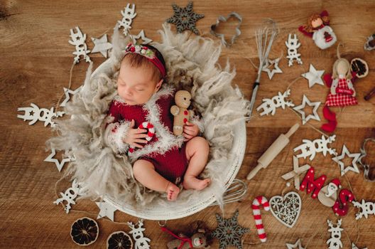 Newborn baby girl sleeping wearing Santa costume and holding knitted ginger man toy lying in basin. Adorable infant child kid napping Christmas portrait