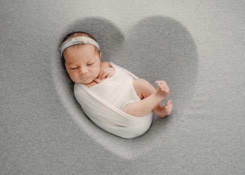 Newborn baby girl sleeping in heart shape form swaddled in white fabric. Adorable infant child kid napping during studio photoshoot