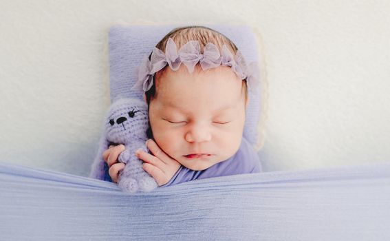 Newborn baby girl holding cute knitted bunny toy and sleeping. Adorable infant child kid napping wearing flower wreath