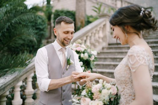 Smiling groom puts the ring on the bride finger while standing on the steps. High quality photo