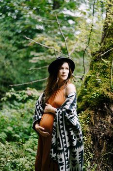 Pregnant woman hugs belly while standing near mossy tree in park. High quality photo