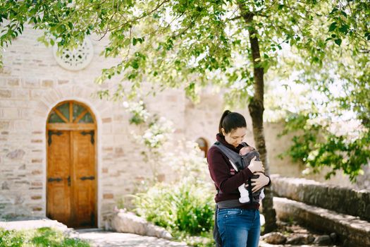 Mom hugs a baby in a sling while standing near a tree in the yard of a stone building. High quality photo