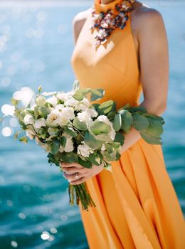 Woman in a yellow dress with a bouquet of flowers stands against a background of water. Close-up. High quality photo