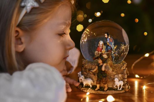 Girl looking at a glass ball with a scene of the birth of Jesus Christ near christmas tree