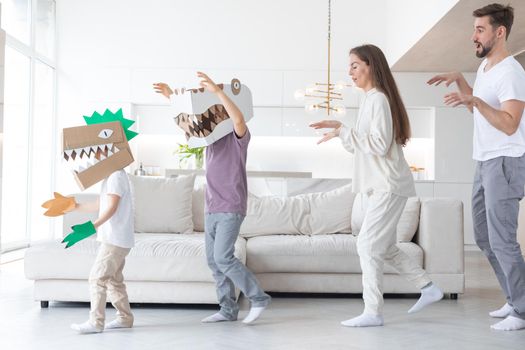 Happy family of parents and two children playing dinosaurs at home, children wearing handmade costume mask of cardboard