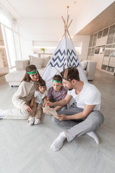 Happy family of parents and two children playing indian at home, wigwam tent, feather roach, reading book about native american indian culture