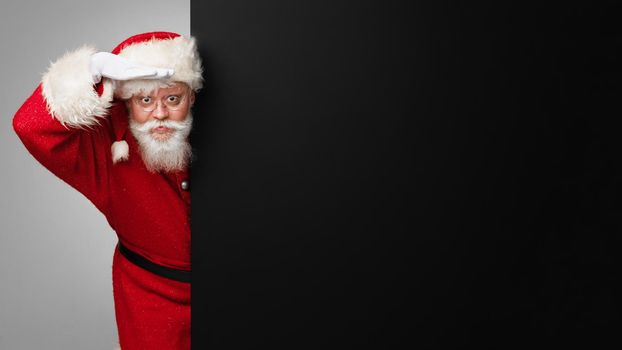 Santa Claus look to future holding hand on forehead standing near black banner with copy space for text