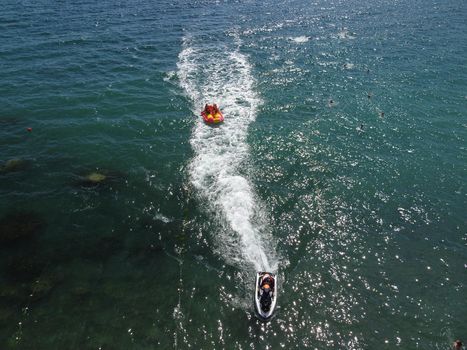 Happy people swim on air mattress behind a high speed water bike. Tourists ride on inflatable watercraft boat. Sea attraction. Water boat skating people on large Inflatable raft. Aerial copter view