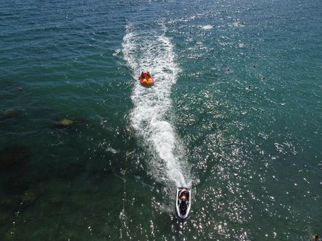 Happy people swim on air mattress behind a high speed water bike. Tourists ride on inflatable watercraft boat. Sea attraction. Water boat skating people on large Inflatable raft. Aerial copter view