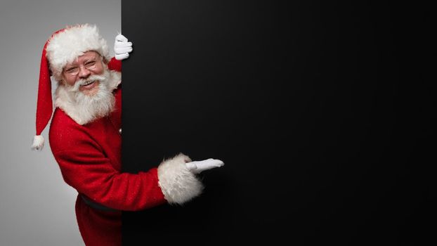 Happy smiling Santa Claus pointing with open hand palm at black banner with copy space for text