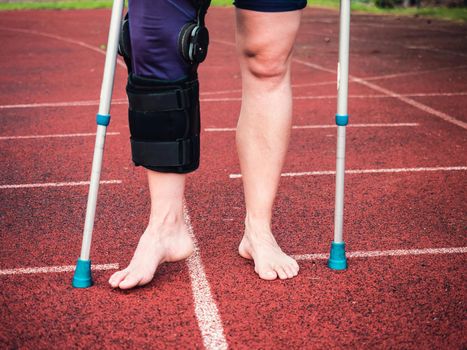 Woman athlete on crutches, wearing a wrist brace and knee support, bandaged leg.  Woman walks on red running surface