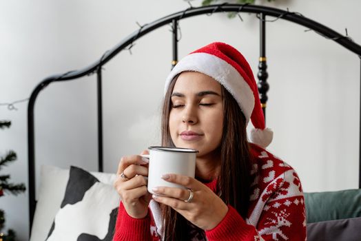 Merry Christmas and Happy New Year. Young brunette woman in red sweater and santa hat sitting on the bed celebrating christmas having cup of hot drink