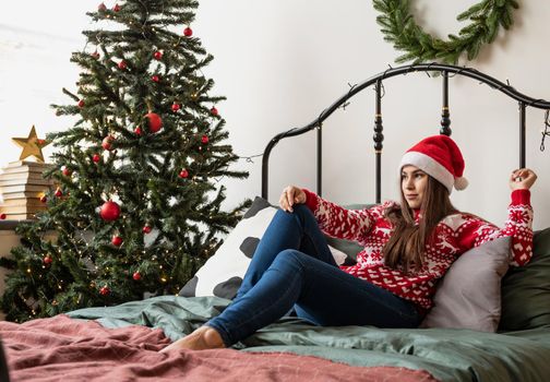 Merry Christmas and Happy New Year. Happy young woman in red sweater and santa hat sitting on the bed celebrating christmas