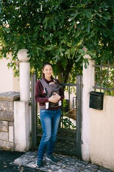 Mother with a baby in a carrier backpack stands near the gate in the yard. High quality photo