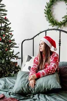 Merry Christmas and Happy New Year. Happy young woman in red sweater and santa hat sitting on the bed celebrating christmas