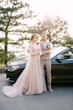 Bride holds groom arm near the convertible on the road. High quality photo