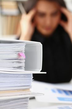 Close-up of tired and exhausted woman looks at documents touch head with hands. Huge pile of document folders on workplace. Headache, low energy concept