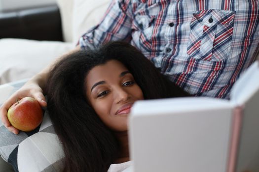 Portrait of latino american young woman reading interesting book laying on fathers lap. Smiling female get new knowledge, spare time at home. Hobby concept