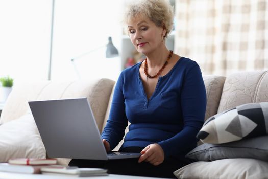 Portrait of senior woman learn how to use modern laptop device at home on sofa. Aging female explore new technology on pastime. Education, hobby concept