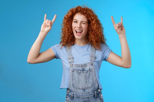 Daring cool sassy redhead ginger girl curly natural hairstyle winking joyfully yelling yeah enjoy awesome party dancing music festival show rock-n-roll heavy metal gesture have fun blue background.