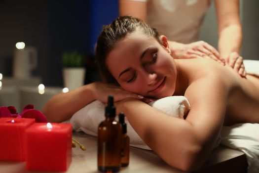 Portrait of young woman enjoying being massaged by professional masseuse master. Chilling female get body treatment in spa, candles and oils. Care concept