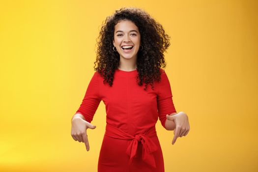 Waist-up shot of assertive charming and happy young woman with curly hairstyle laughing joyfully, smiling and pointing down as showing cool place to check out over yellow background. Advertisement concept
