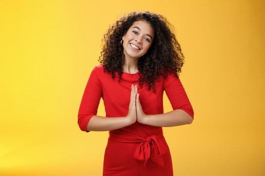 Welcome come inside. Portrait of friendly and polite good-looking female host in red dress with curly hair holding hands in namaste gesture tilting head and smiling as inviting guests in asian style.