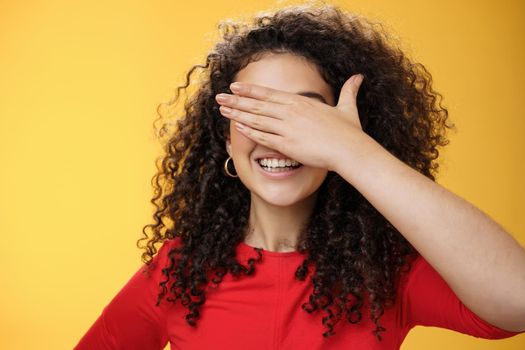 Close-up shot of dreamy happy young cute woman with curly hair covering eyes with palm as counting or playing peekaboo smiling broadly anticipating surprise happen over yellow background.