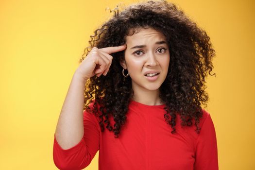 Are you crazy. Portrait of frustrated and confused woman with curly hair holding index finger near temple and raising eyebrows shocked being annoyed with stupid actions of weirdo over yellow wall.