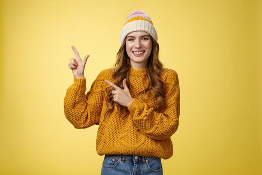 Charming carefree smiling young 20s woman wearing hat sweater pointing upper left corner sideways sharing interesting link awesome promotion delighted recommend cool product, yellow background.