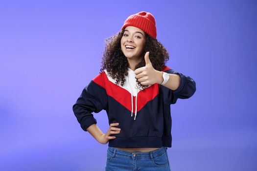 Nice work, keep up. Delighted and supportive charming sportswoman in winter beanie, sweatshirt and watch showing thumbs up with satisfied smile, giving approval, liking and cheering great effort. Emotions and body language concept