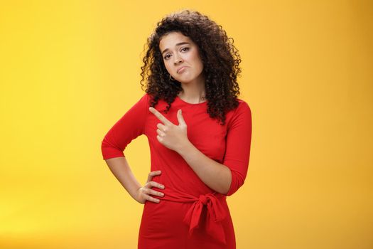 Upset disappointed and unimpressed attractive young european curly-haired woman in red dress pursing lips in gloomy face, frowning displeased as pointing at upper left corner from regret and dislike.