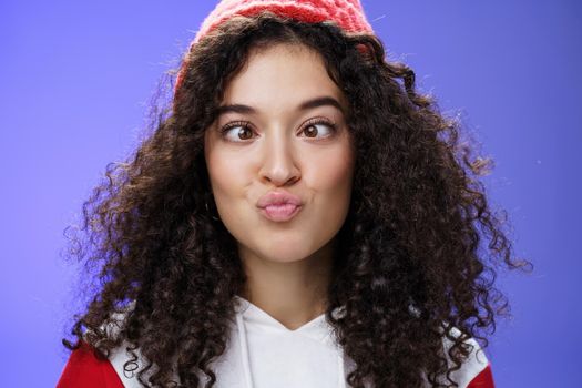 Headshot of funny and playful girl fooling around making faces not scared being funny folding lips like duck and squinting ice, aping standing childish and hilarious over blue wall in winter hat.