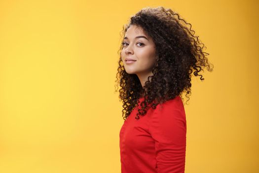 Profile shot of stylish and cool attractive curly-haired european 25s woman in red dress turning at camera and smiling broadly with delighted carefree expression, having fun over yellow background.