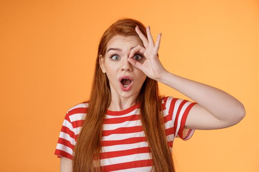 Surprised excited shocked young redhead girl gasping impressed look through okay circle, stare camera astonished awesome sale, open mouth say wow thrill and amazement, orange background.