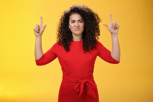 Displeased, unimpressed moody girlfriend with curly hairstyle in red elegant dress raising hands pointing up and grimacing unsatisfied and disgusted expressing dislike and averstion over yellow wall.