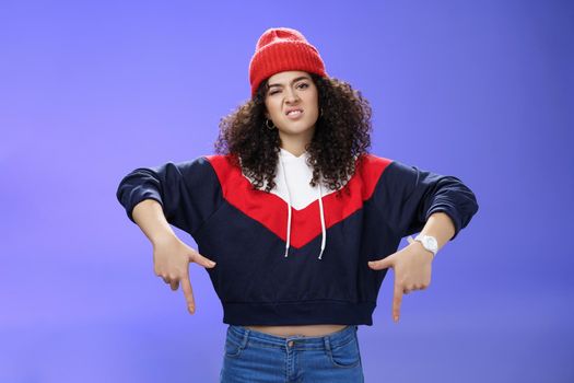 Snobbish and displeased stylish and cool street girl with curly hair in beanie and sweatshirt pointing down grimacing as feeling unsatisfied and disappointed posing against blue background.