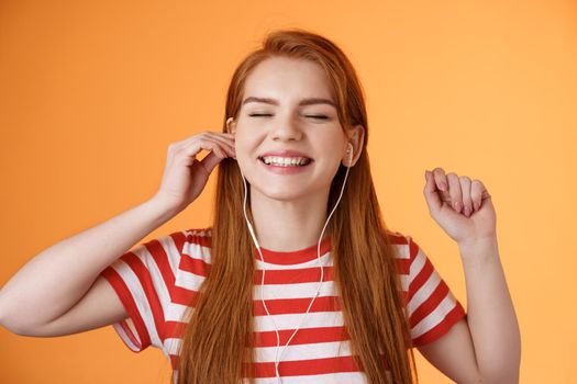Carefree happy redhead woman put earphone smiling delighted, listen favorite song, close eyes relaxed, enjoy good music playlist, dancing joyfully, satisfied awesome headphones quality.