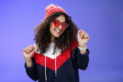 Stylish and feminine girlfriend dancing having fun feeling upbeat as liking weather feeling warm and cozy in stylish red beanie and sunglasses dancing with raised hands and broad smile over blue wall.
