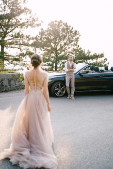 Bride stands across the street from groom leaning on the convertible. High quality photo