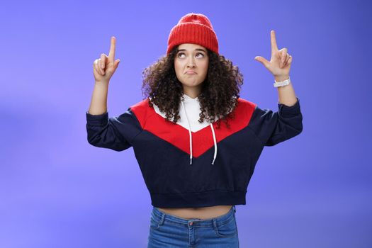 Girl feeling disappointed and gloomy as looking at bad weather making sad smile, pursing lips down raising hands staring up upset and unsatisfied standing displeased against blue background.