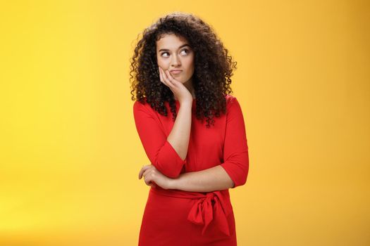 Waist-up shot of cute girlfriend scheming feeling bothered with some idea or thought smirking looking right interested and impatient as making up plan standing thoughtful over yellow background. Copy space