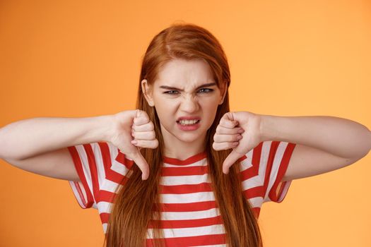 Displeased unhappy redhead female viewer frowning disappointed, give bad feedback watch awful movie, show thumbs-down hateful grimace, complain bad service, dislike show, orange background.