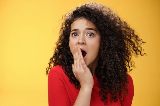 Close-up shot of concerned and shocked worried woman with curly hairstyle open mouth wide and covering it with palm popping eyes at camera impressed and upset over yellow background. Emotions and faciale xpressions concept